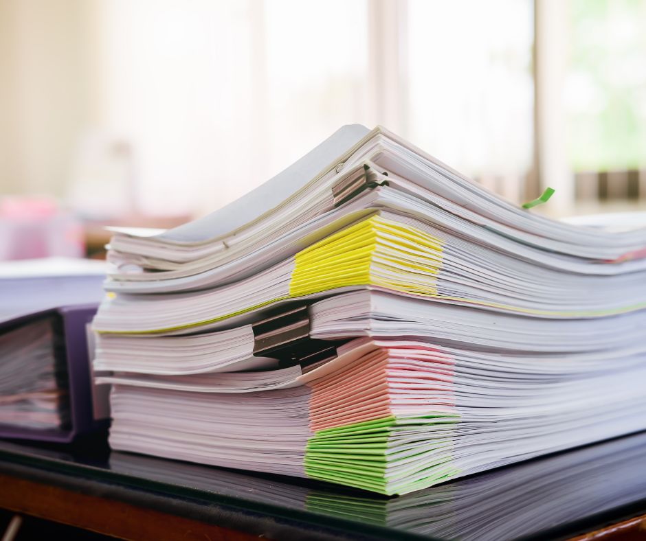 A large stack of organized documents sits on a desk. The papers are clipped together with metal binder clips and highlighted with fluorescent sticky notes in various colors. Sunlight filters through a window in the background, illuminating the workspace.