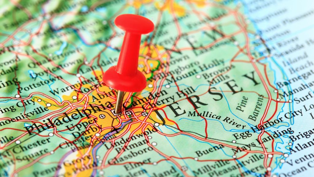A red pushpin marks a location on a detailed map, highlighting the area between Philadelphia and New Jersey. The map features roads and city names.