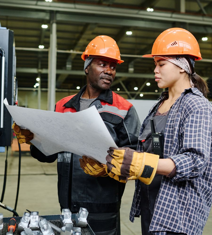 Two workers wearing hard hats review a blueprint inside a manufacturing facility.