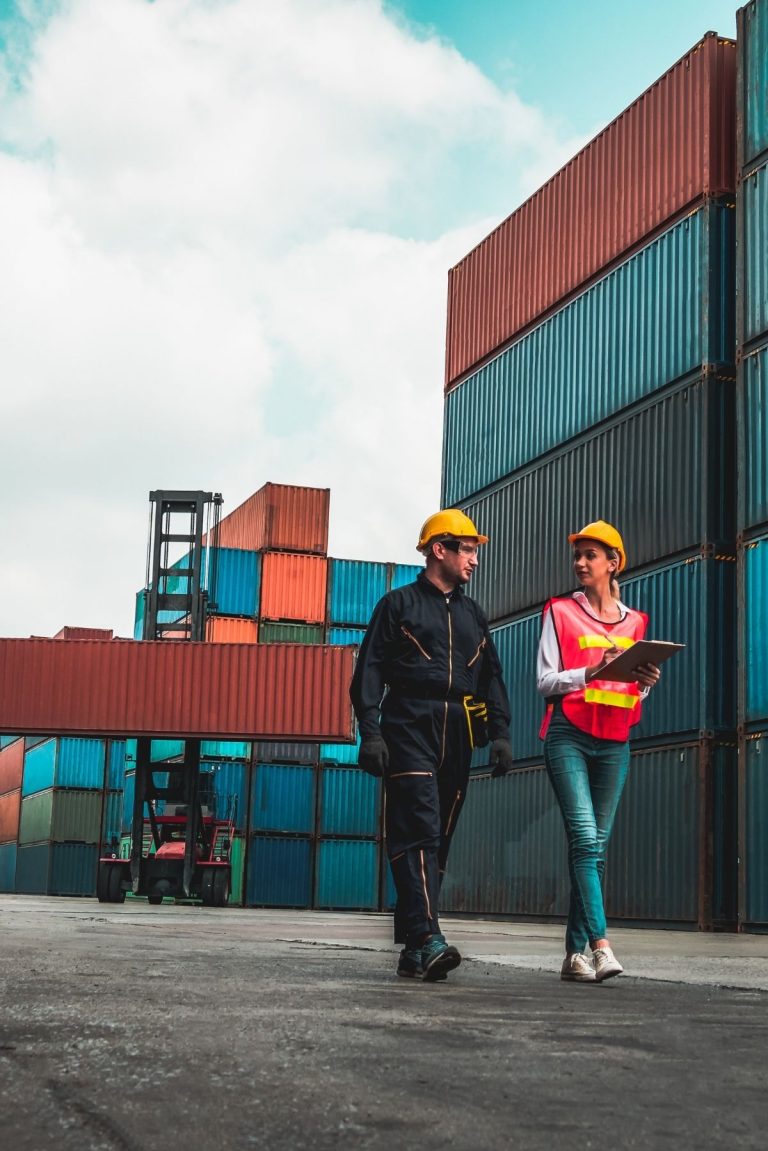 Two workers walking through a logistics yard talking and taking notes