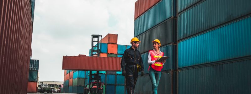 Two workers walking through a logistics yard talking and taking notes