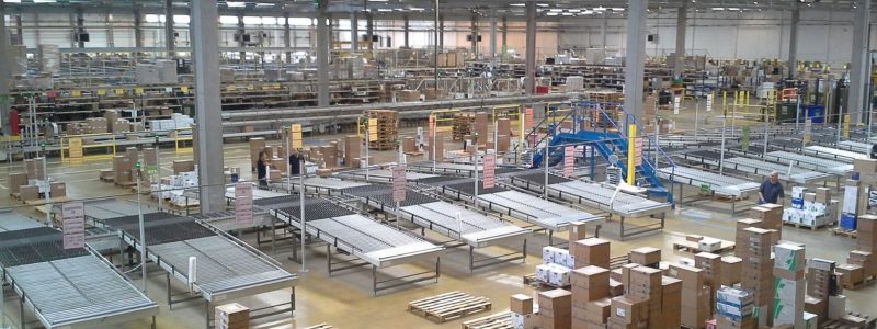 Warehouse floor with conveyor belts, stacked boxes, and pallets inside a large manufacturing facility.